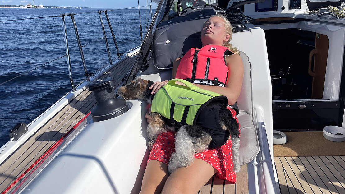 帆船主人的女儿和狗穿着救生衣在帆船的甲板上休息，狗也穿着救生衣。