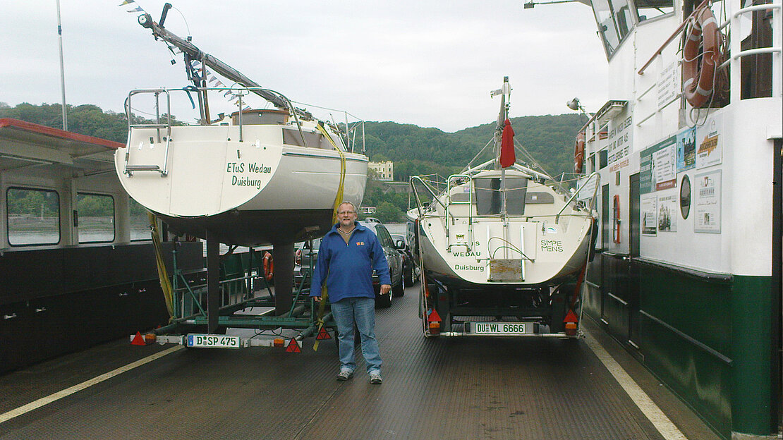 A man stands next to a sleek boat, trailered aboard a ferry.