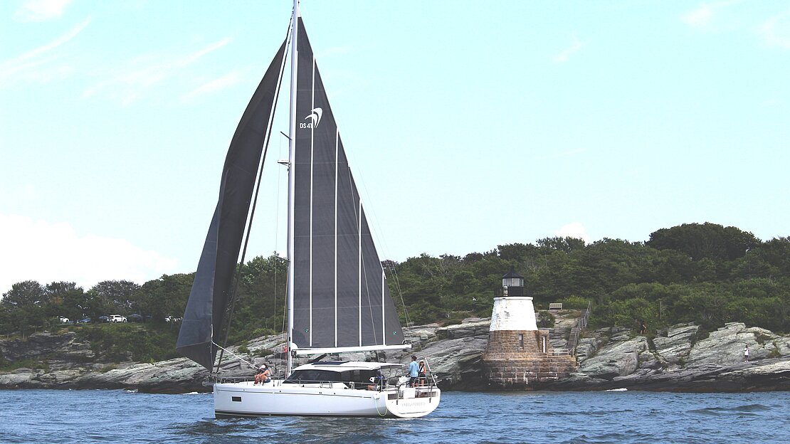 Championship of Luxury Blue Water Yachts, sailboat sailing past lighthouse on rocky shore, blue sky with white clouds.